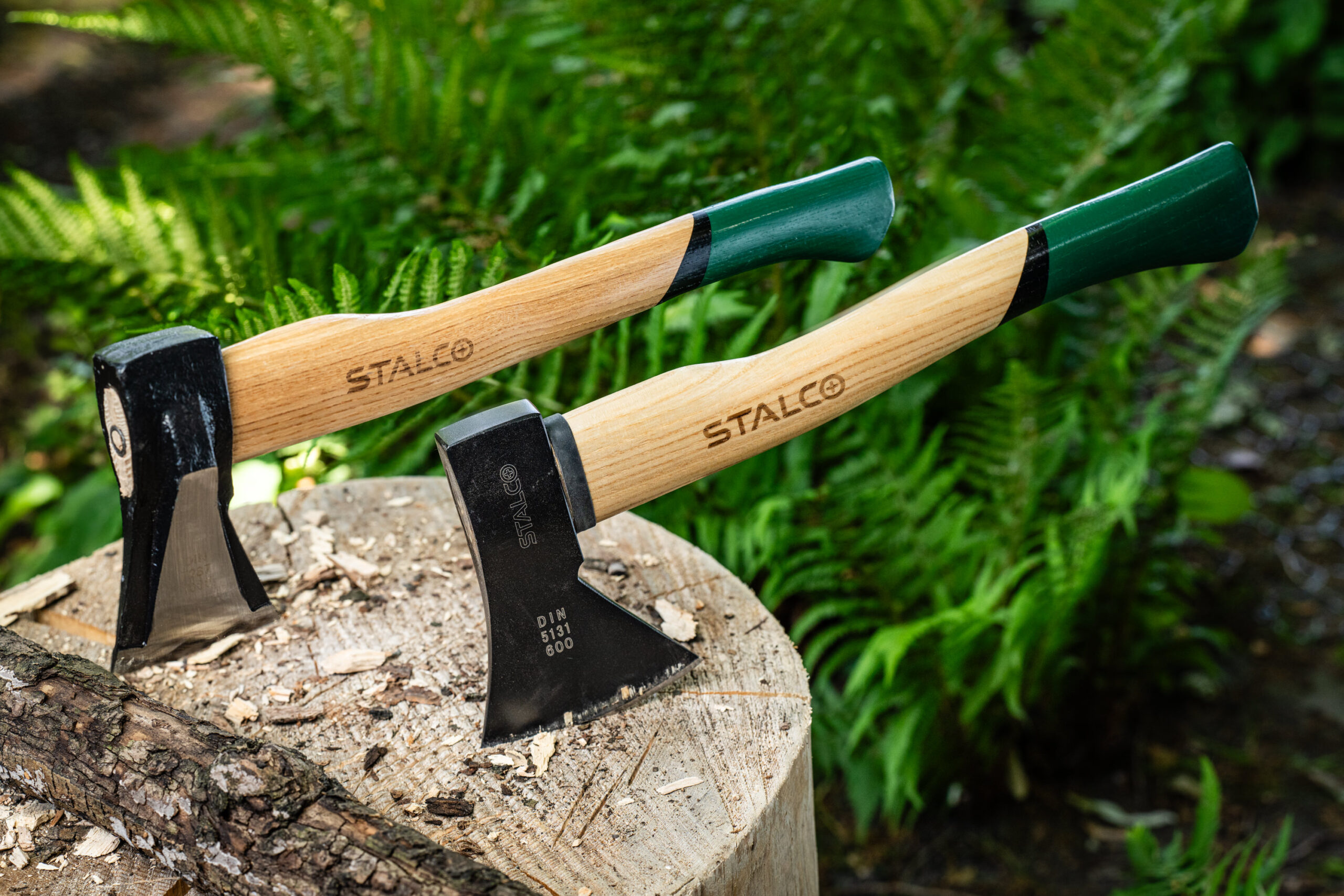Dve sekery Stalco na pni v lese: sekera 600g DIN 5131 so sklolaminátovým poriskom a štiepacia sekera 2kg s dreveným poriskom a klinovou hlavicou.